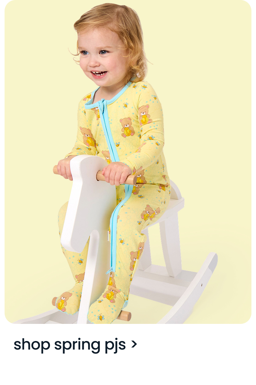 "Child wearing a yellow zip‑front footed pajama with teddy bear prints, sitting on a white rocking toy. Text below reads ‘shop spring pjs.’"