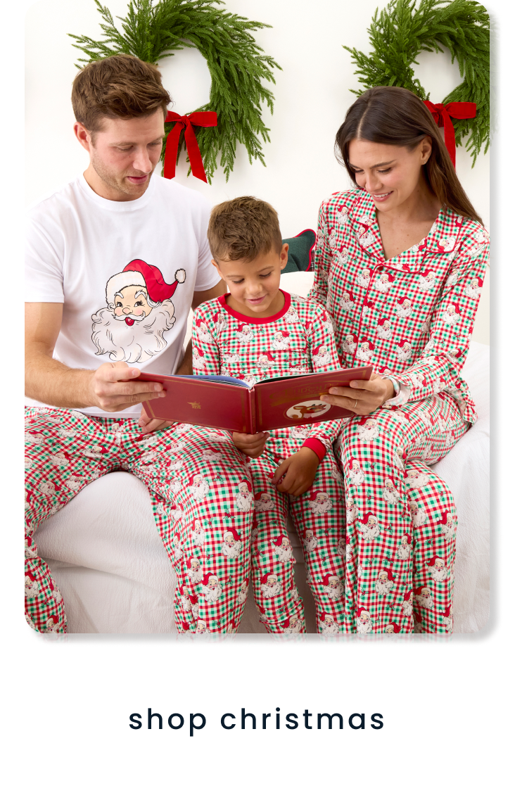 "Family wearing matching Santa-print holiday pajamas while reading a book in front of wreath decorations. Text below reads ‘shop christmas.’"