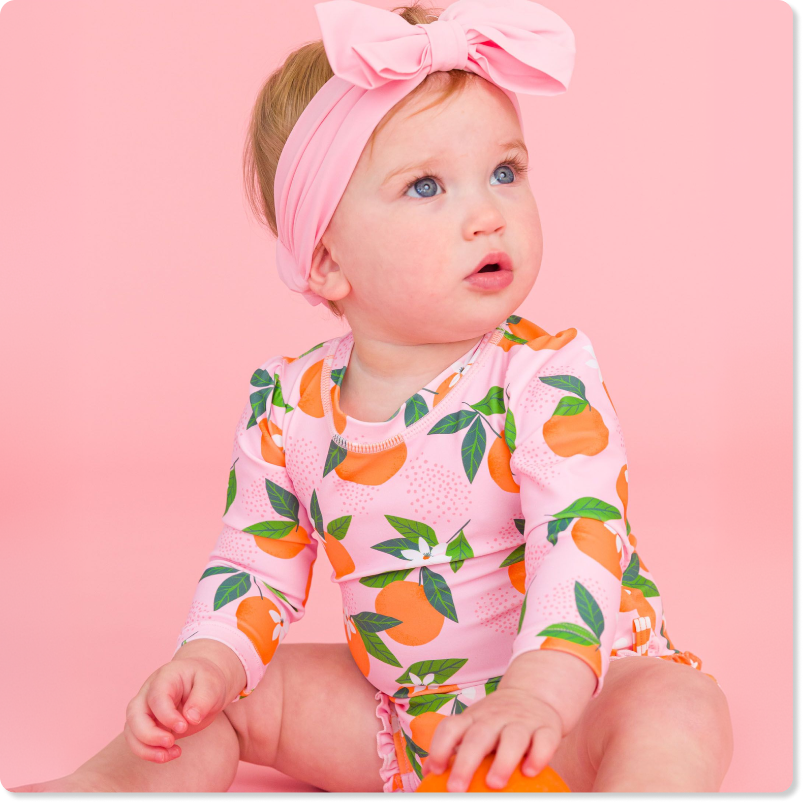 "Baby wearing a pink citrus-print long-sleeve swimsuit and pink bow headband, seated against a pink background and holding an orange."