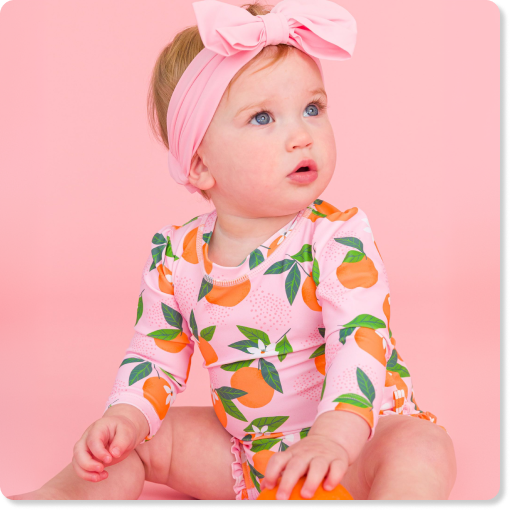 "Baby wearing a blue-and-white striped long-sleeve swimsuit, light blue sun hat, and heart-shaped sunglasses while standing in shallow pool water beside a blue float."