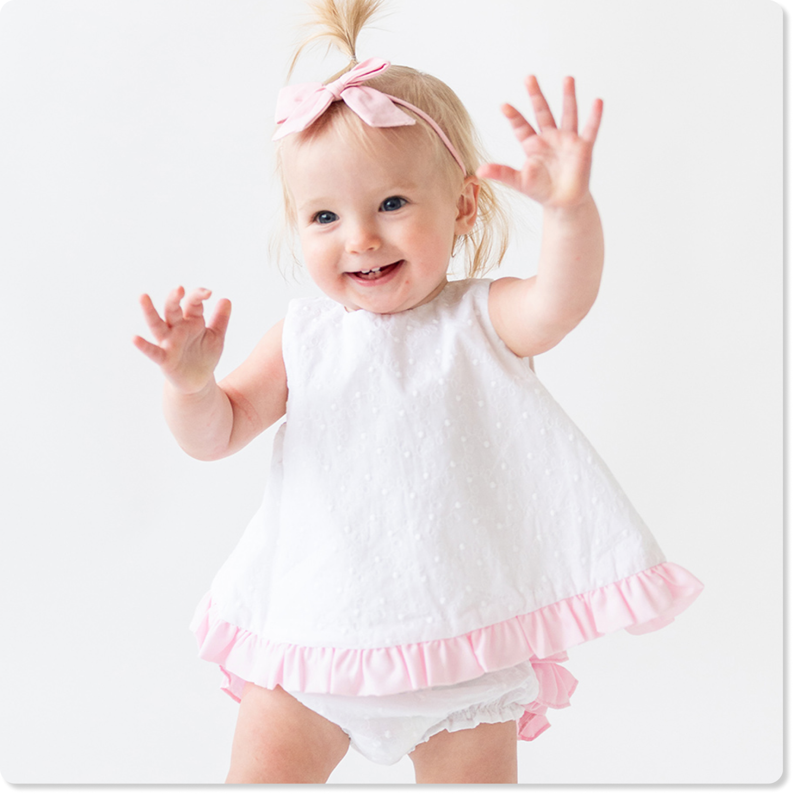 "Baby wearing a white sleeveless dress with pink ruffle trim and matching bloomers, paired with a pink bow headband on a light background."