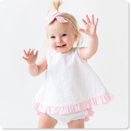 "Baby wearing a white sleeveless dress with pink ruffle trim and matching bloomers, paired with a pink bow headband on a light background."