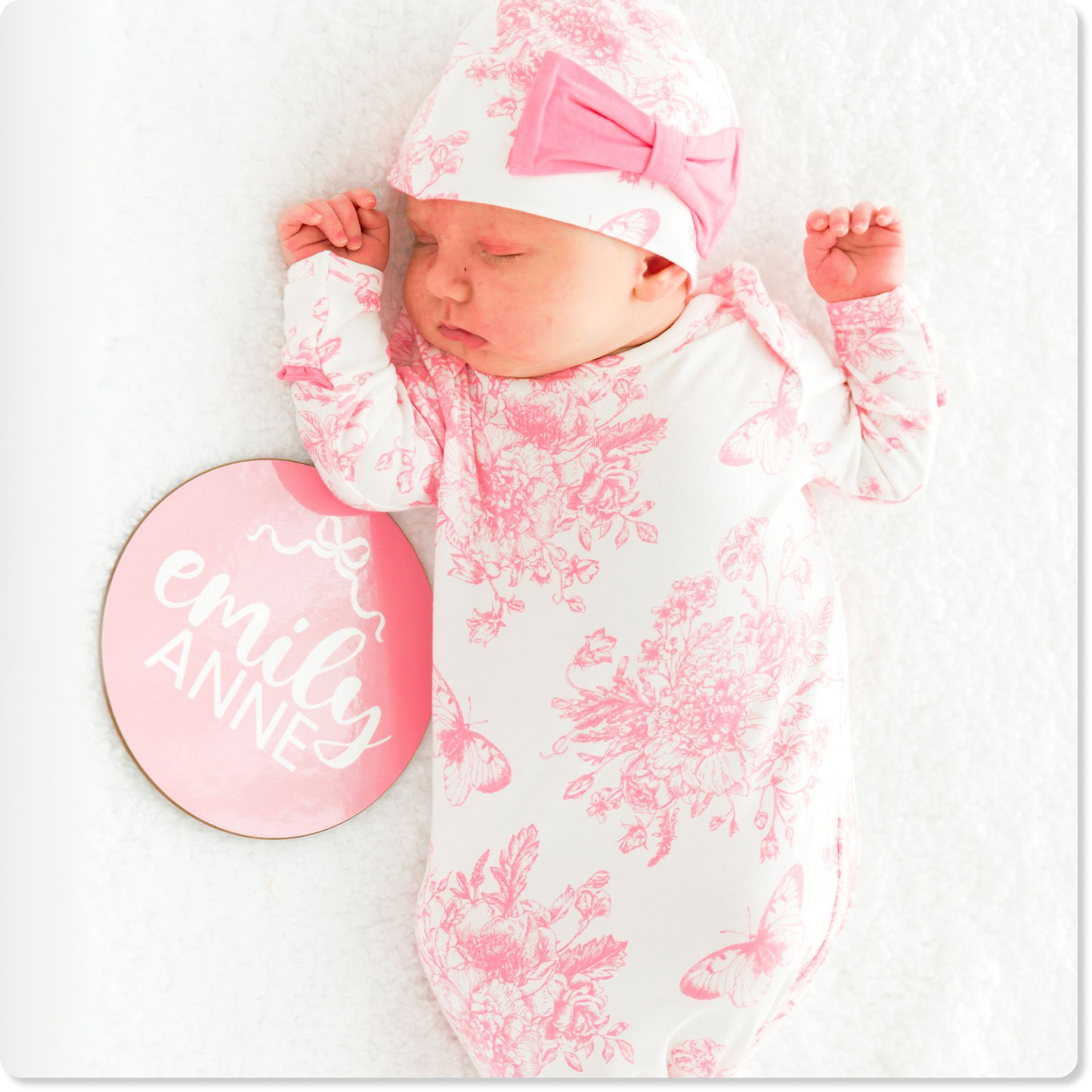 "Newborn wearing a white gown with pink floral and butterfly print and a matching hat with a pink bow. A milestone sign beside the baby reads &lsquo;Emiley Anne.&rsquo;"