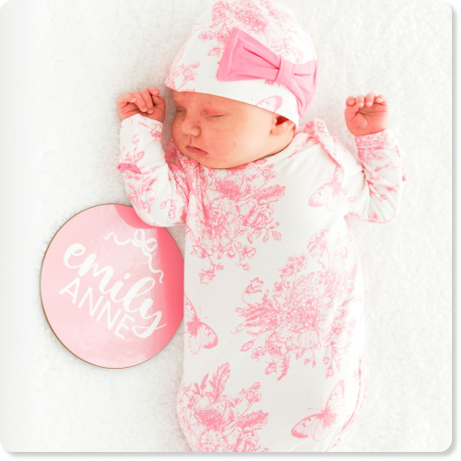 "Newborn wearing a white gown with pink floral and butterfly print and a matching hat with a pink bow. A milestone sign beside the baby reads &lsquo;Emiley Anne.&rsquo;"