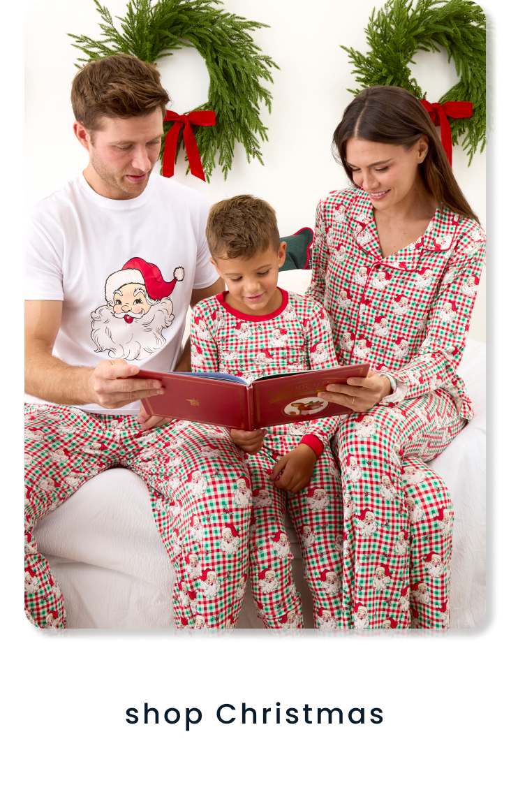 "Family wearing matching Santa-print holiday pajamas while reading a book in front of wreath decorations. Text below reads ‘shop christmas.’"
