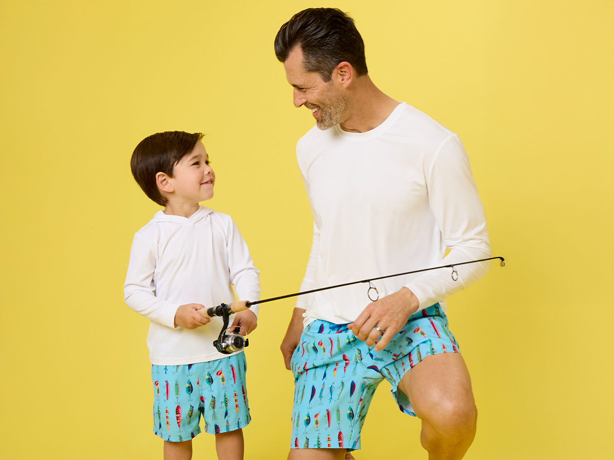 Adult male and child wearing matching turquoise swim trunks with fishing lure prints, holding fishing rods against a yellow background. 