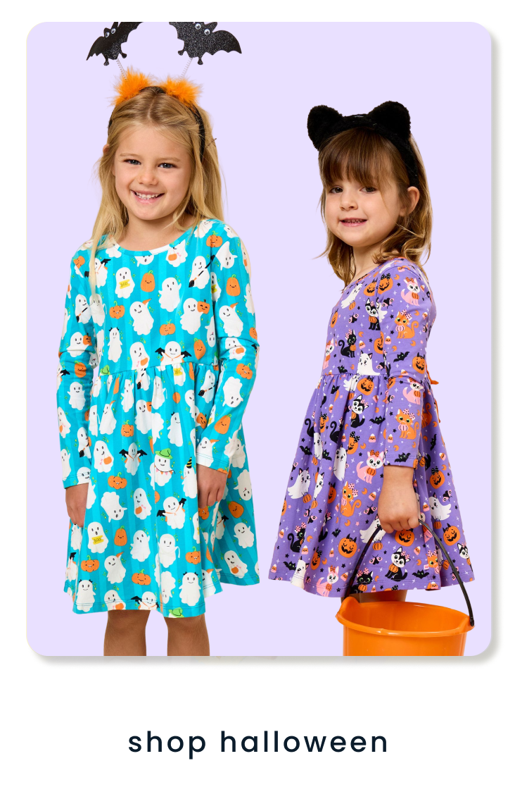 "Two children wearing Halloween-themed dresses with ghost, pumpkin, cat, and bat prints. One holds an orange trick-or-treat bucket. Text below reads ‘shop halloween.’"