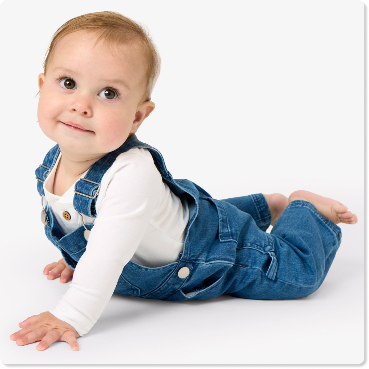 "Baby wearing denim overalls layered over a white long-sleeve bodysuit, posed on a light gray background."