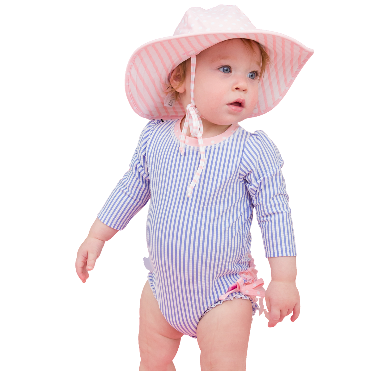 "Baby wearing a blue-and-white striped long-sleeve swimsuit and pink-striped sun hat on a pastel pink background. Swimsuit features pink bow details at the leg openings."